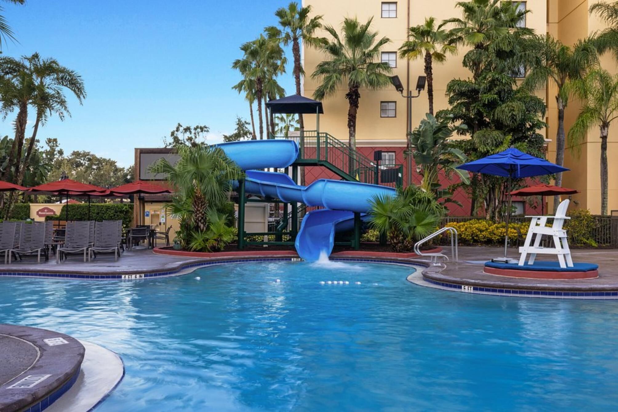 Une piscine avec un toboggan bleu, entourée de palmiers et bordée d’un bâtiment doté de balcons.