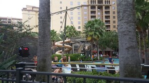 Zona de piscina en un resort, con tobogán de agua, palmeras y un edificio de varios pisos al fondo.