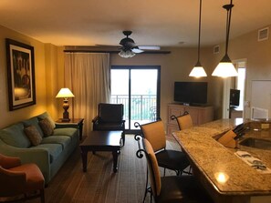 Habitación de hotel con sofá, mesa de comedor, sillas, cocina con mostrador, ventilador de techo y televisor.