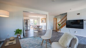A modern living room with a flat-screen TV, a dining area with a table and chairs, a staircase, and a kitchen with a refrigerator. - Luxury Family Getaway with Designer Touches (Atlantic City)