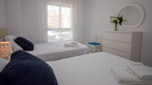 Ein Hotelzimmer mit zwei Betten, einem Fenster mit durchsichtigen Vorhängen, einem weißen Schminktisch mit Spiegel und einer Vase mit Blumen auf dem Schminktisch.