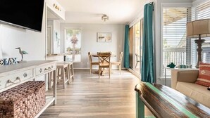Amplia sala de estar con suelo de madera, una mesa de comedor con sillas, un sofá, un televisor montado en la pared y una ventana con cortinas.