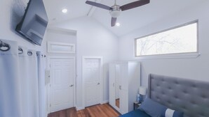 Ein Schlafzimmer mit Bett, Deckenventilator, an der Wand montiertem Fernseher und einem Fenster mit Blick auf Bäume.