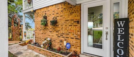 Un mur extérieur en briques, agrémenté d’une pancarte de bienvenue, d’une porte et d’un petit jardin où poussent des courges.