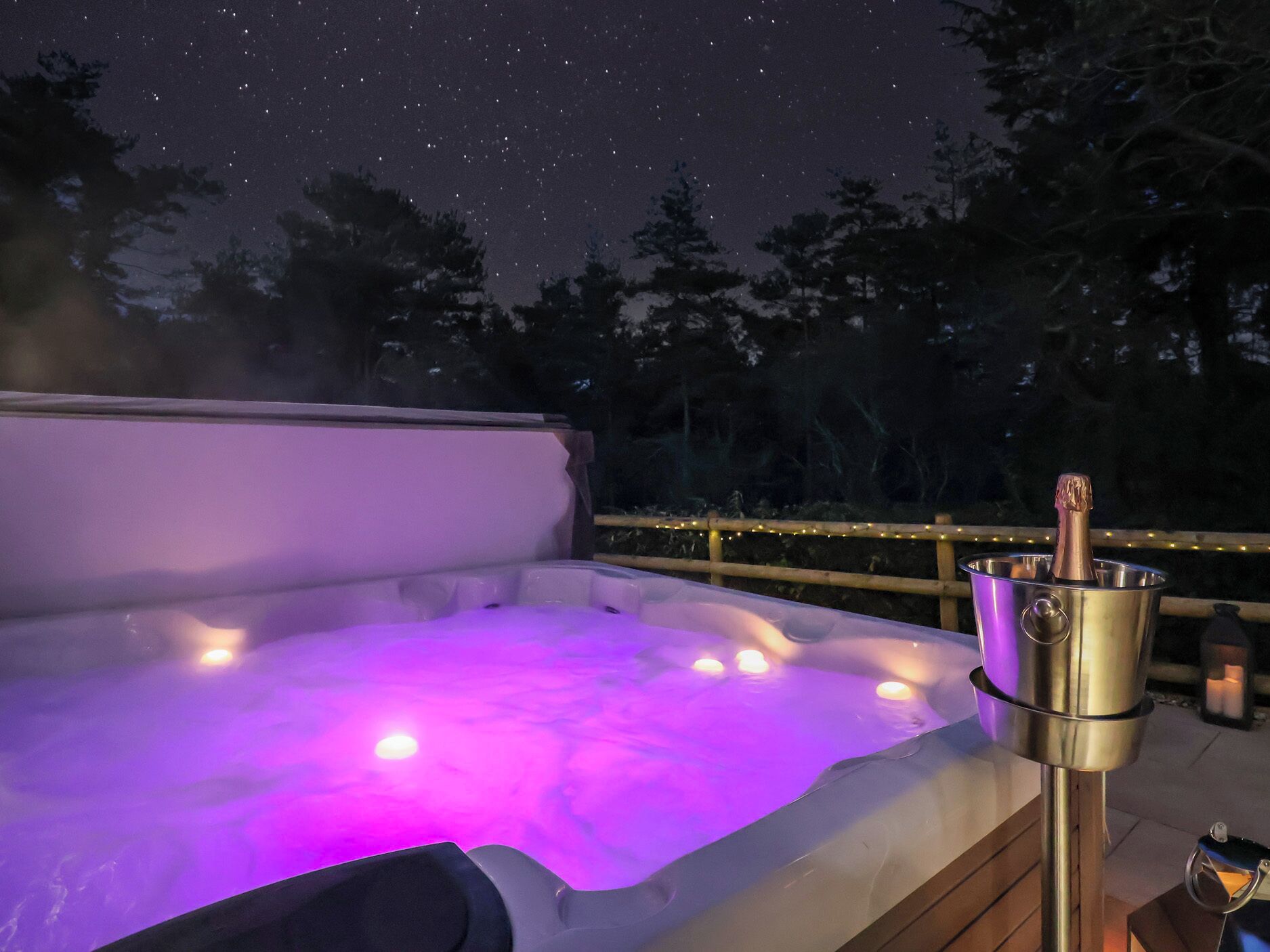 A hot tub with a metallic bucket containing a bottle, surrounded by a wooden deck and string lights, under a starry night sky.
