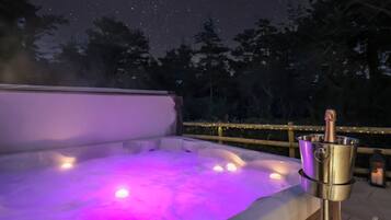 A hot tub with a metallic bucket containing a bottle, surrounded by a wooden deck and string lights, under a starry night sky.