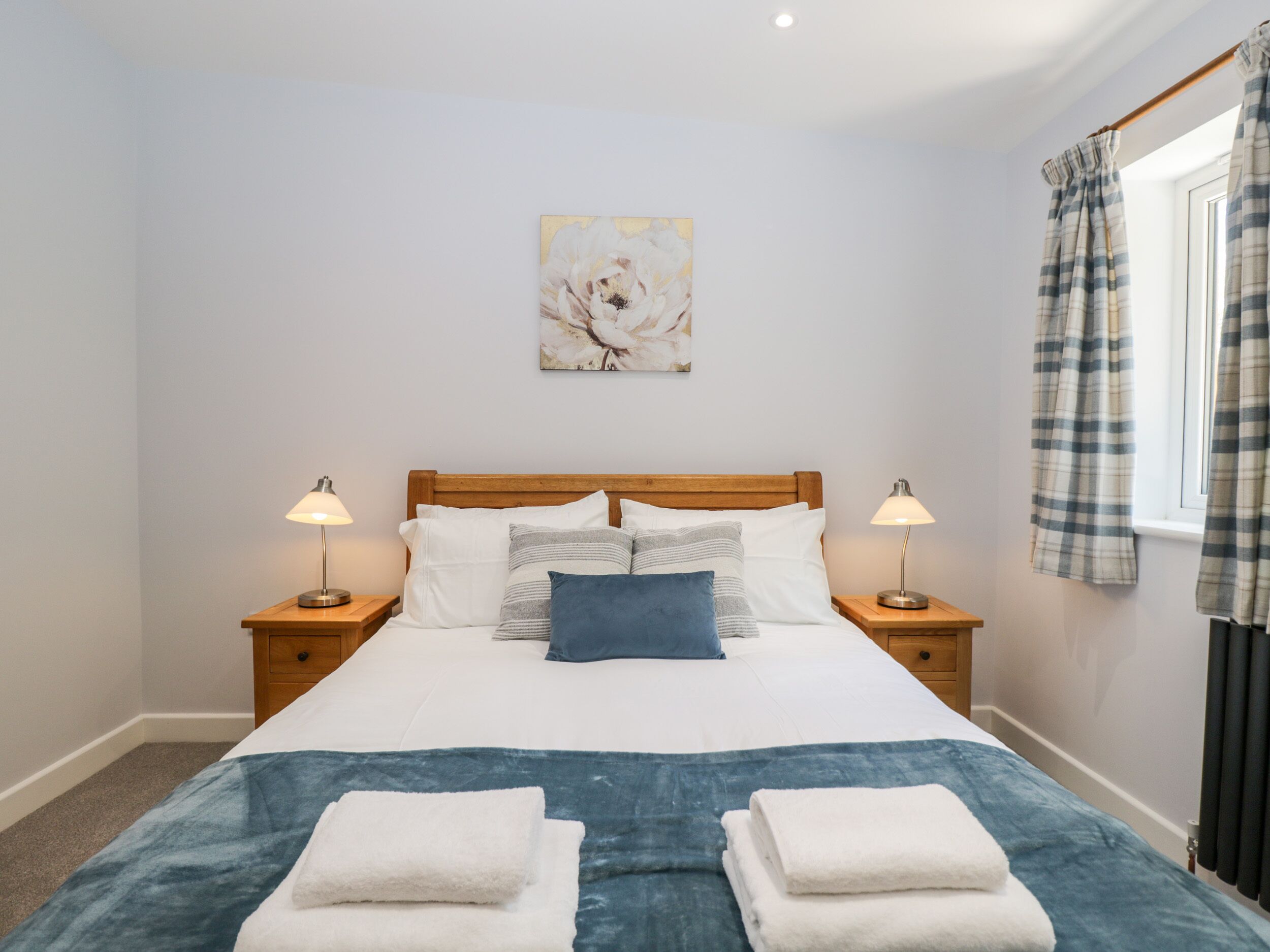 A neatly made bed with white and blue bedding, two bedside lamps, and a window with plaid curtains.