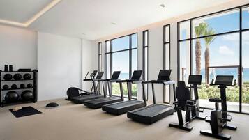 A modern gym with treadmills, stationary bikes, and a weight rack. Large windows offer a view of palm trees and the ocean.