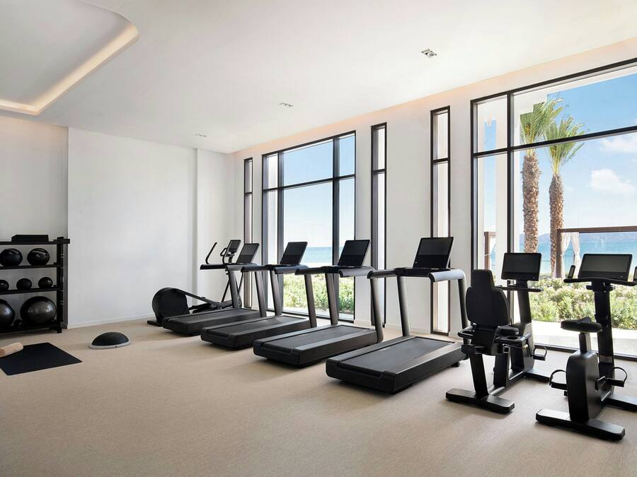 A modern gym with treadmills, stationary bikes, and a weight rack. Large windows offer a view of palm trees and the ocean.