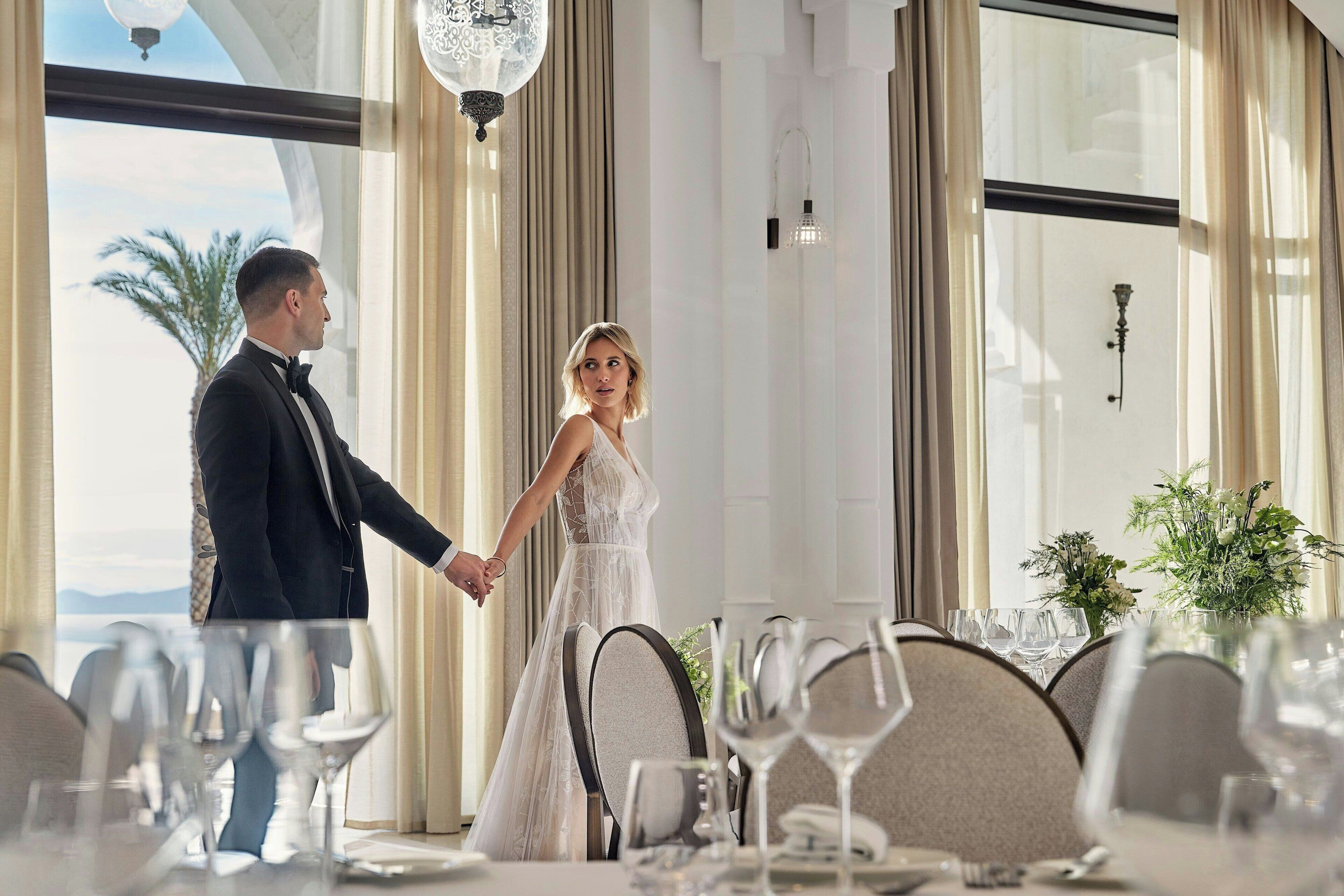Un couple se tenant la main dans un cadre formel, autour d’une table à manger, avec vue sur l’extérieur.