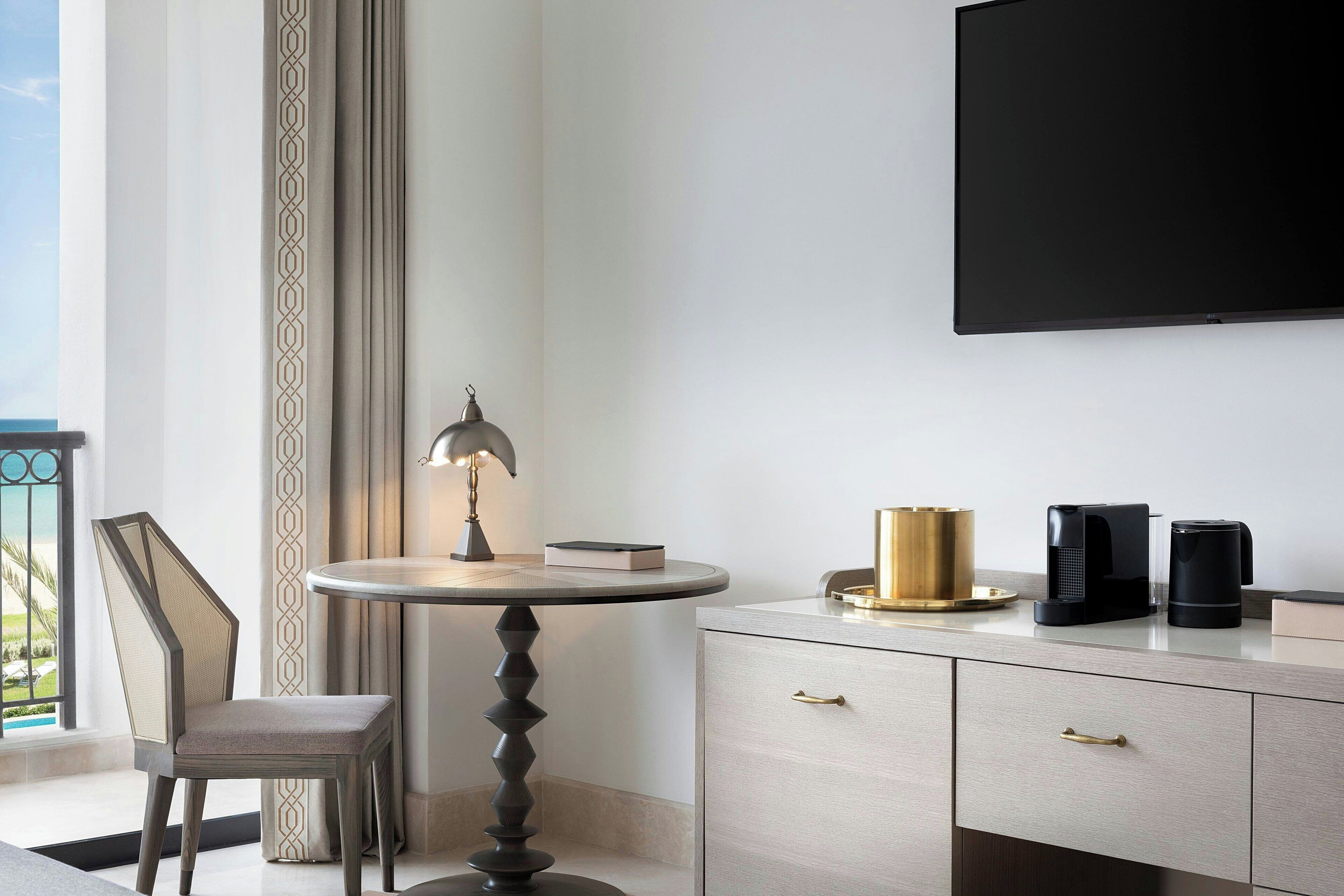 Een moderne hotelkamer met een ronde tafel, een stoel, een aan de muur bevestigde televisie en een minibars met koffiezetapparaten.