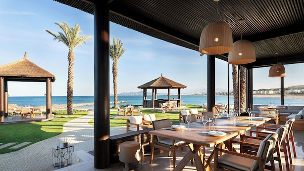 Un área de comedor al aire libre con mesas y sillas, un pabellón con techo de paja y vista al mar.