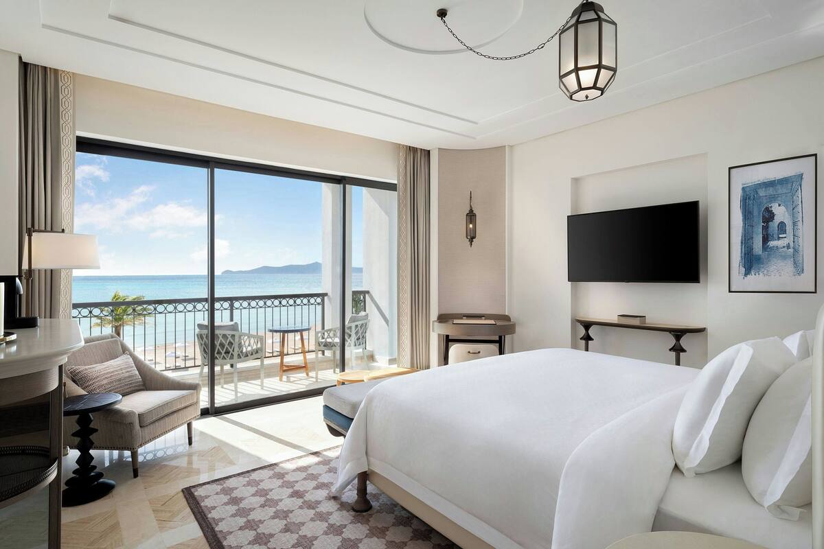 Une chambre d’hôtel avec un grand lit, un bureau avec une lampe, une télévision et un balcon donnant sur la mer.