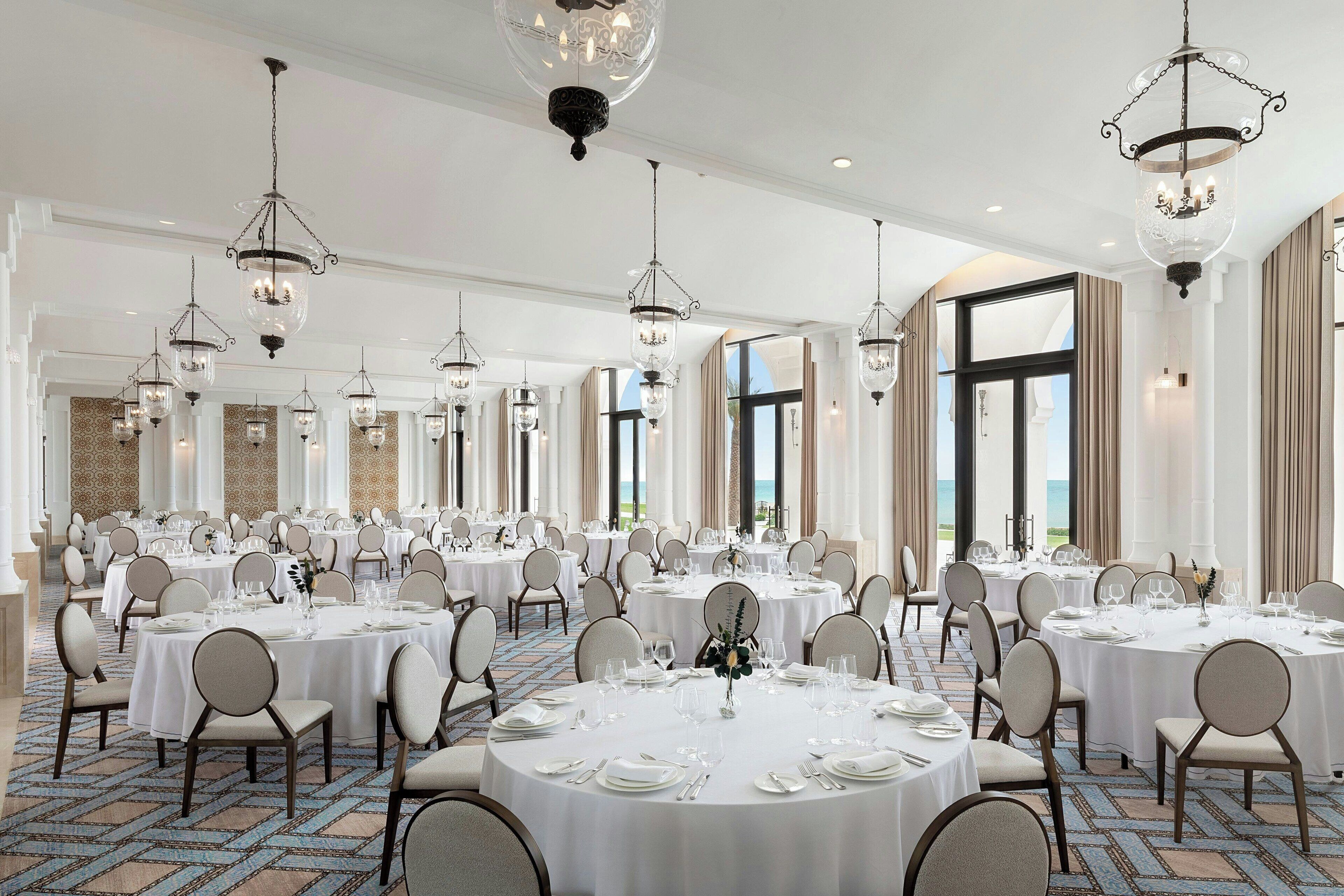 A large banquet hall with round tables set for dining, elegant chandeliers, and a view of the ocean through large windows.