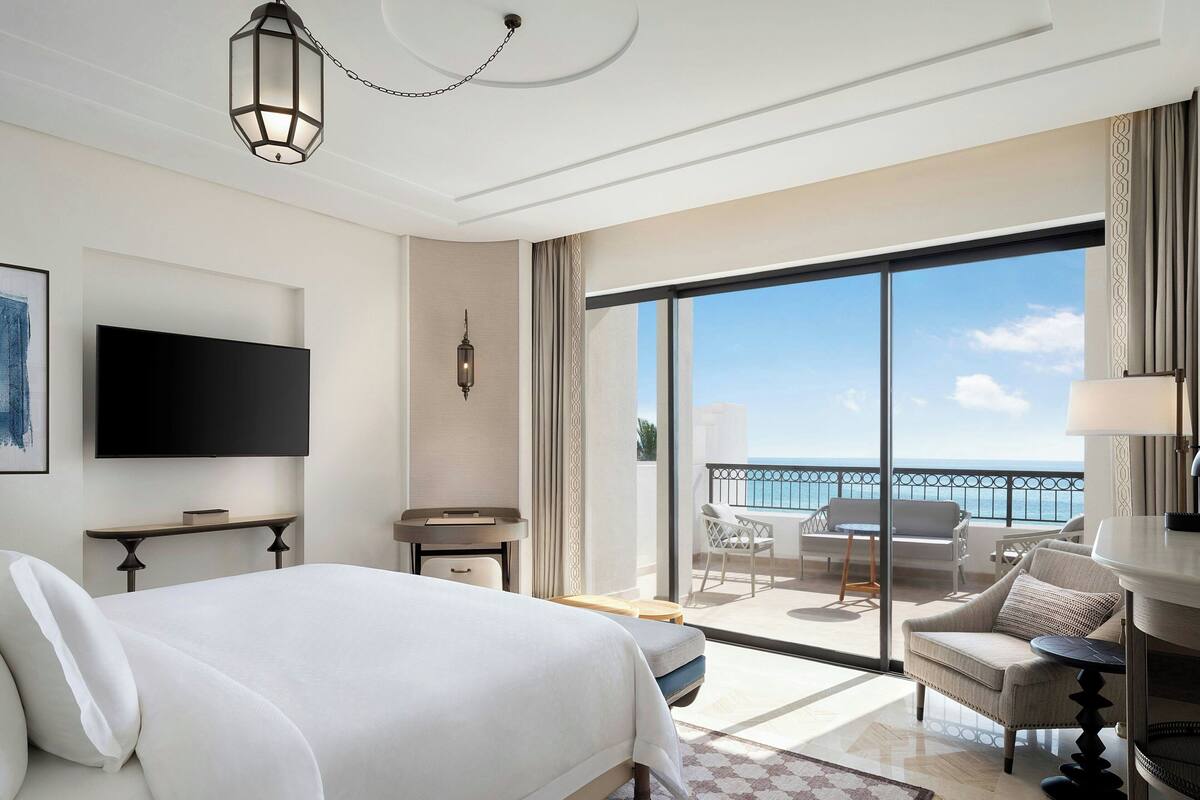Une chambre d’hôtel dotée d’un grand lit, d’une télévision, d’un balcon avec vue sur la mer et d’un coin repas avec une table et des chaises.