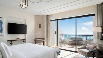 A hotel room with a large bed, a TV, a balcony with a view of the sea, and a dining area with a table and chairs.