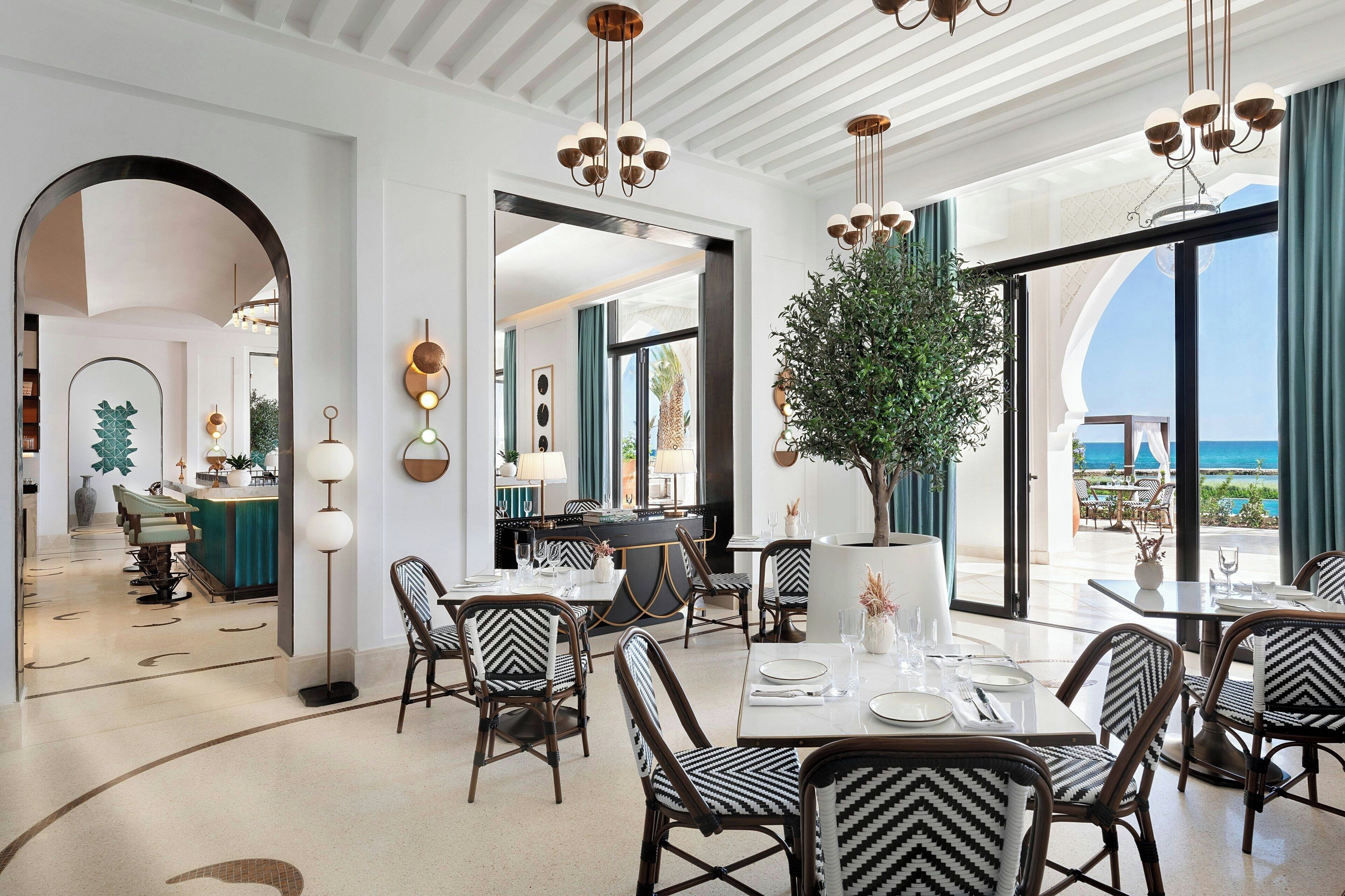 A spacious dining area with modern furniture, large windows offering a view of the ocean, and a central potted plant.