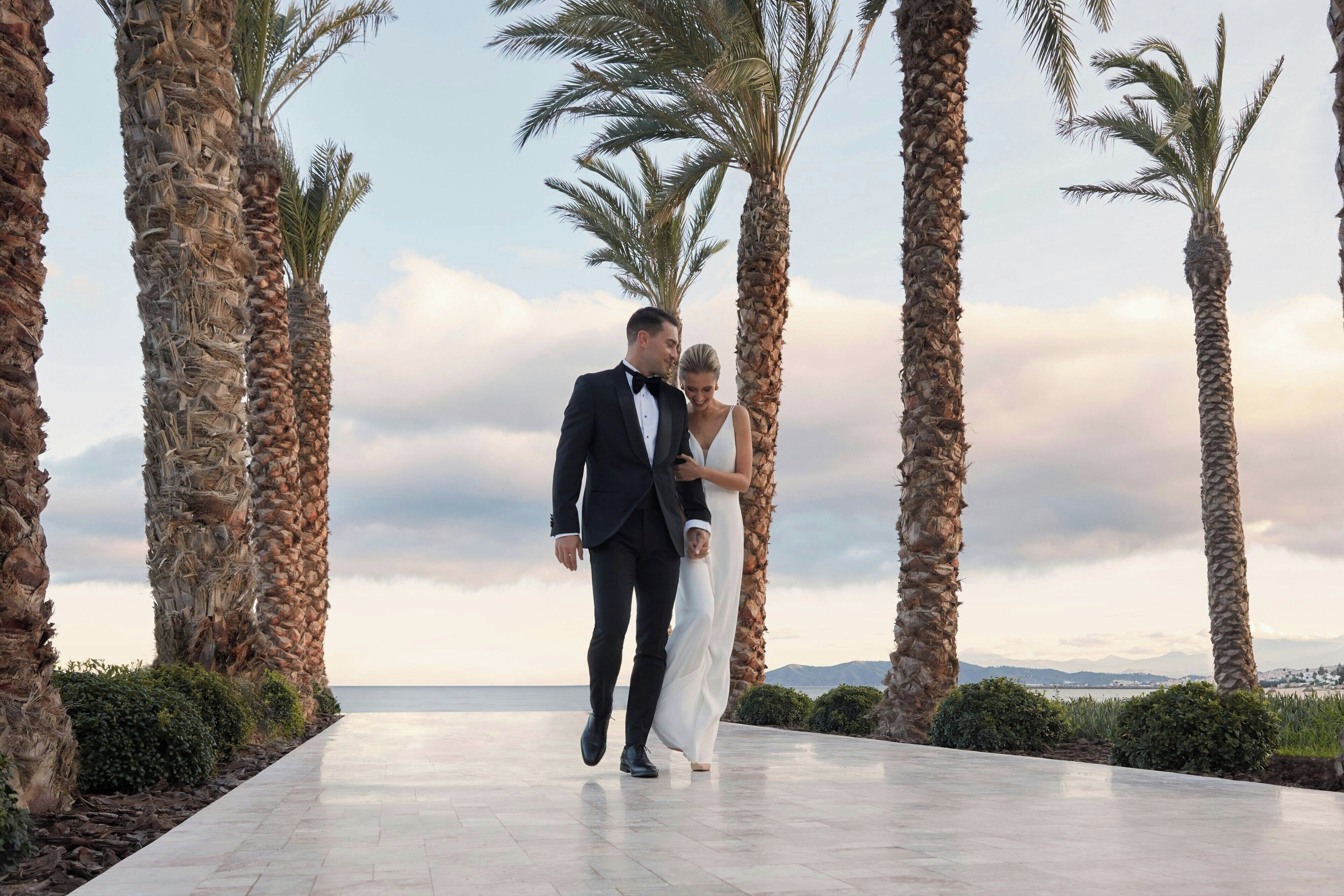 A couple walking on a paved pathway flanked by tall palm trees, with a view of the sea and distant mountains in the background.