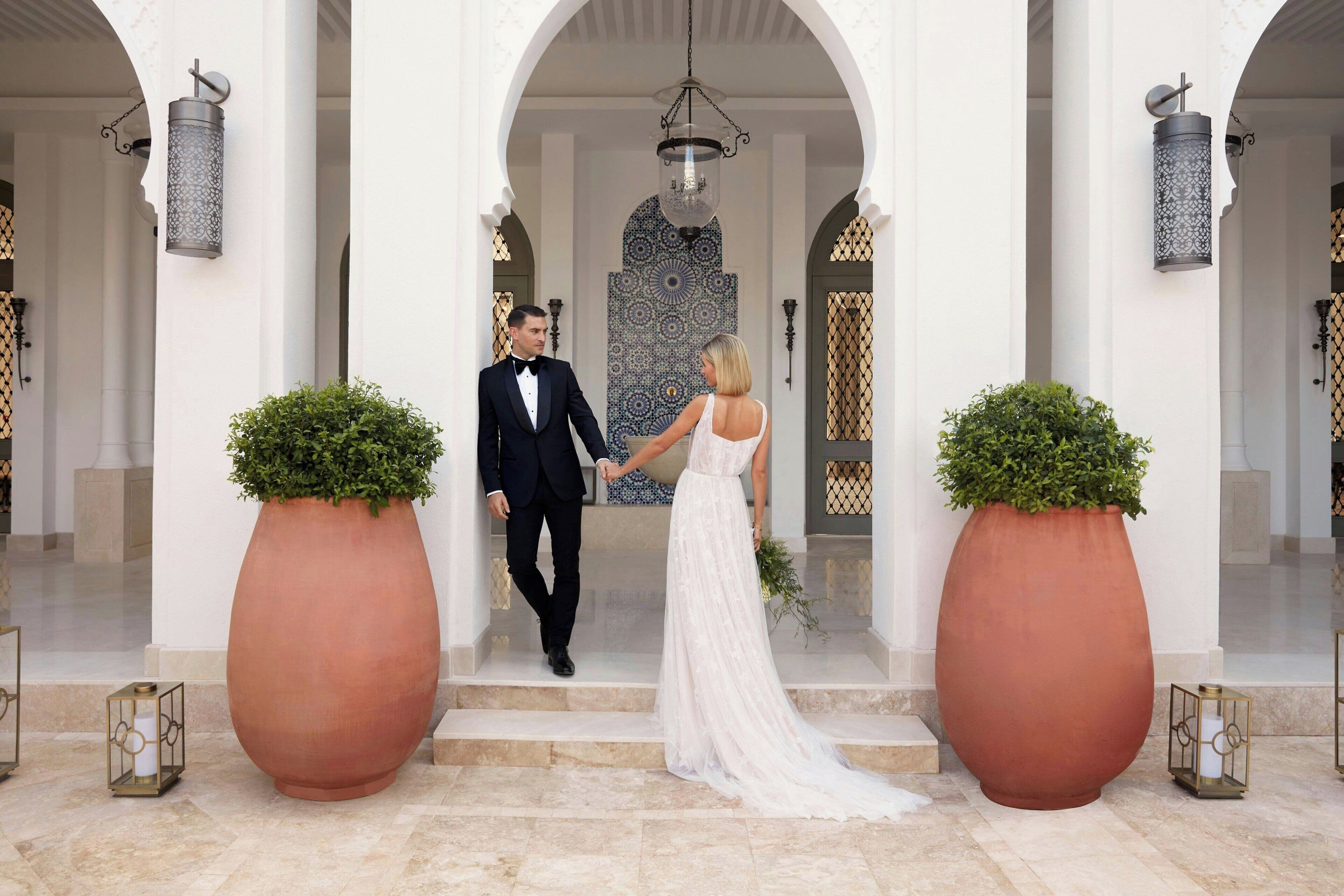 Una pareja caminando de la mano por una escalera de mármol en el vestíbulo de un hotel, con grandes macetas y apliques ornamentales.