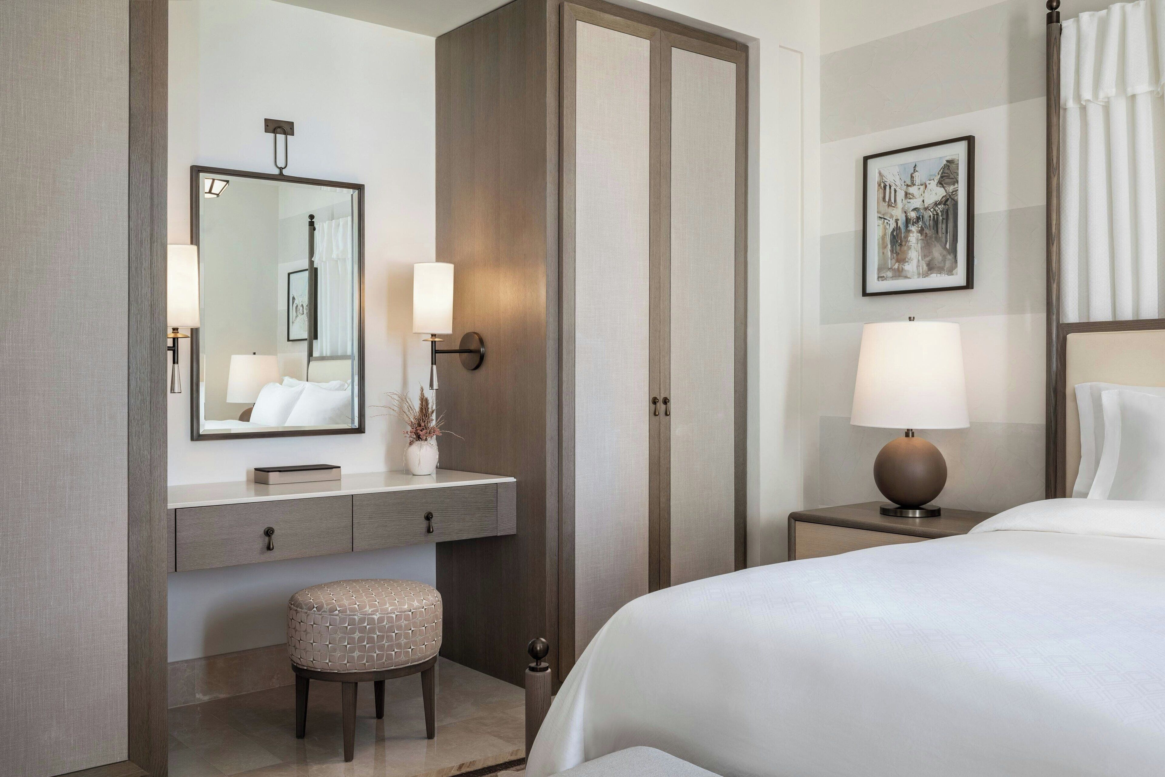 A hotel room with a bed, a nightstand with a lamp, a vanity with a mirror, a small stool, and a door.