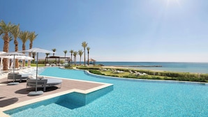 Piscina con sillas de descanso y sombrillas, vista a la playa y palmeras.