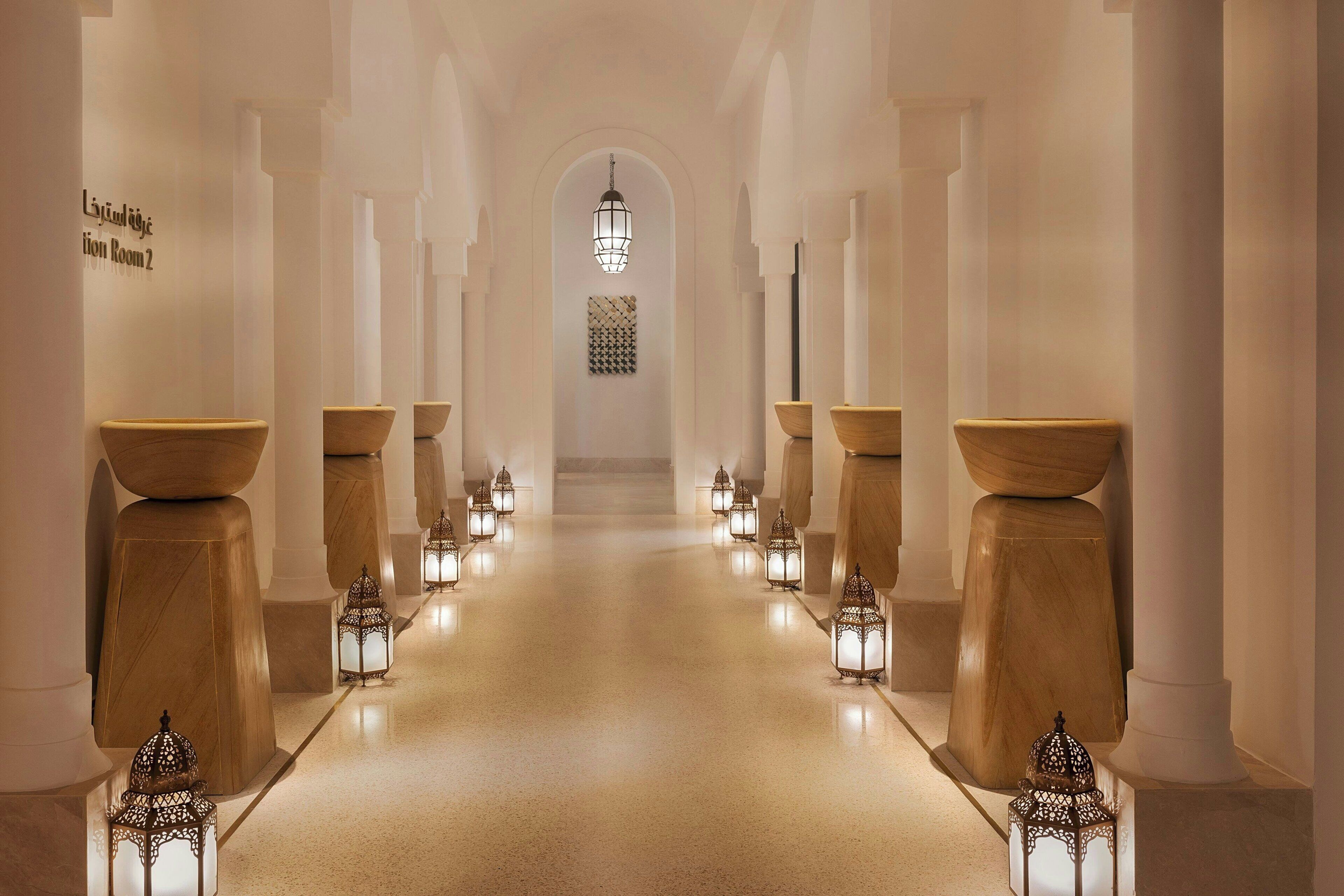 A corridor with a central walkway flanked by columns and lanterns, leading to a large arched doorway.