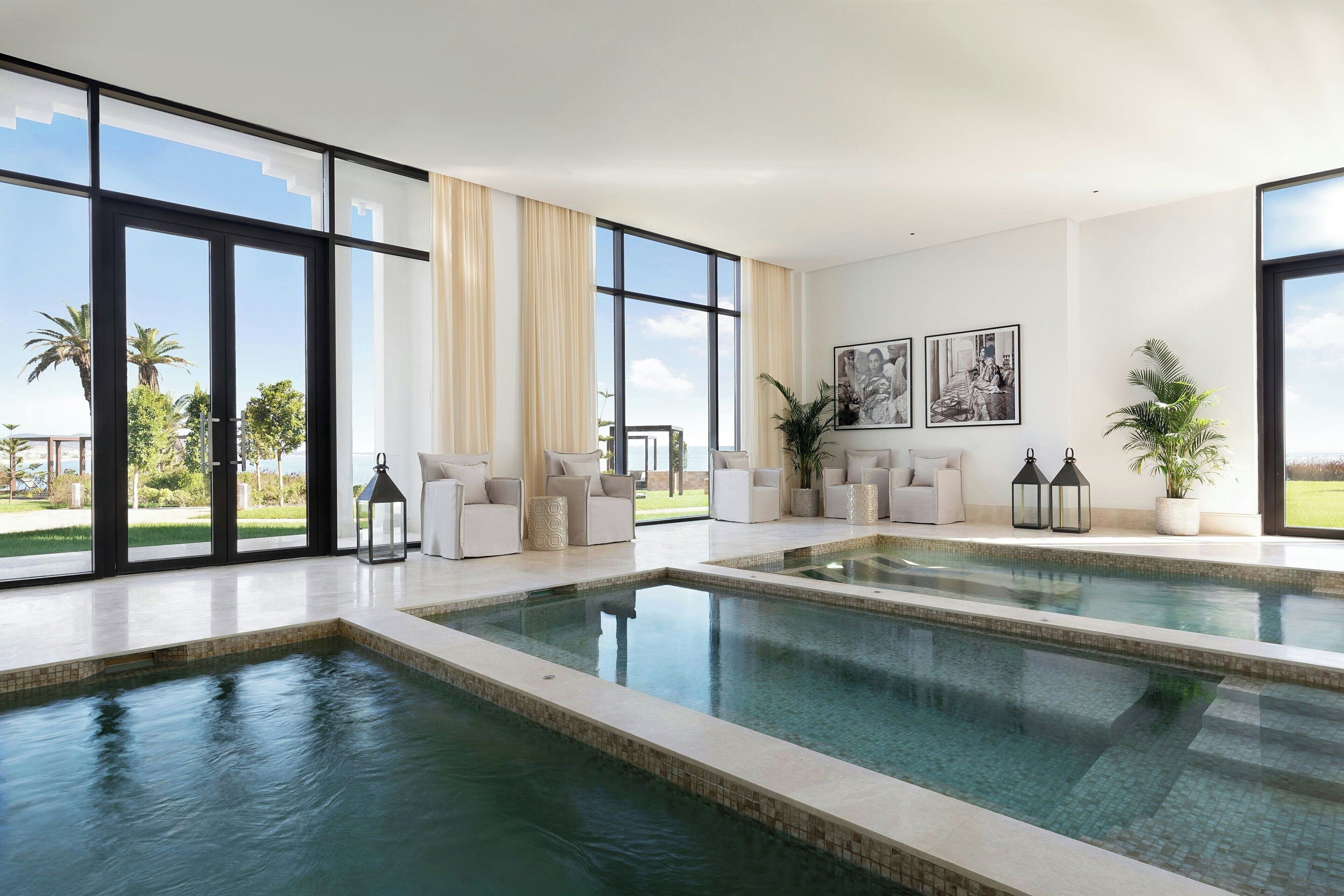 Piscina interior moderna com grandes janelas, área de estar com sofás brancos e vista para o exterior.