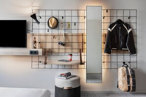 A modern room with a mounted TV, a wooden shelf with a hat, sunglasses, and a camera, and a hanging jacket. - Moxy Vienna City East (Vienna)