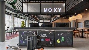 Ein modernes Interieur mit einer Rezeption mit der Aufschrift "MOXY", einer Tafel mit den Hinweisen "Check In" und "Play", einem Skateboard und einem Koffer.