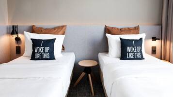 A hotel room with two beds, each with a pillow that reads "WOKE UP LIKE THIS." There is a small round table between the beds, and the room has a neutral color scheme with a gray wall and a wooden floor.