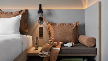 Una habitación de hotel moderna con una cama, una mesita de noche con lámpara, una pequeña mesa con una botella y auriculares, y un sofá con cojines.