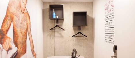 A room with a wall-mounted ironing board, a small shelf with a bottle and a hanger, and a poster with text.