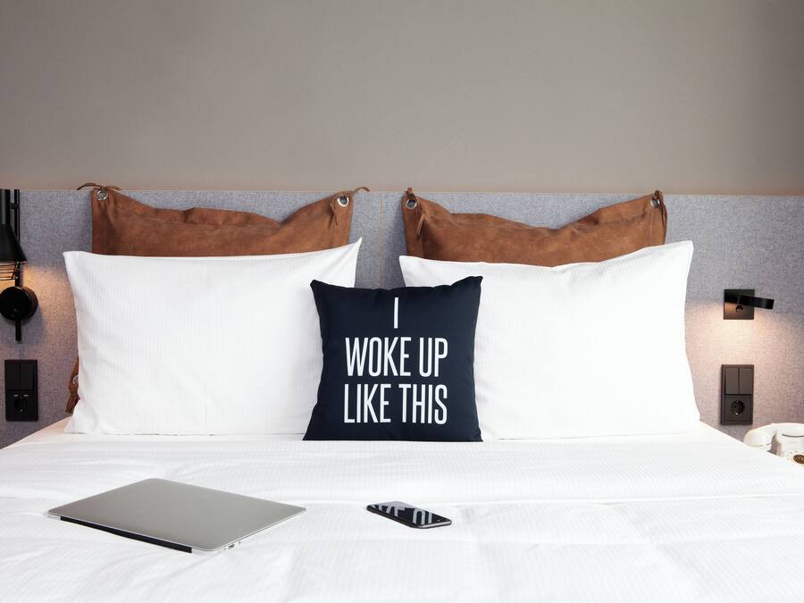 A neatly made bed with a laptop and a smartphone on it, a pillow with the text "I woke up like this," and a telephone on the side.