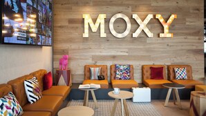 Ein stilvoller Lounge-Bereich mit einem großen, beleuchteten 'MOXY'-Schild, bequemen Sitzgelegenheiten und einer digitalen Wand.