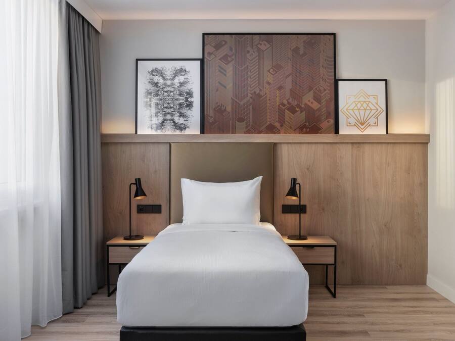 Una habitación de hotel moderna con una cama grande, cabecero de madera, dos mesitas de noche con lámparas y tres obras de arte enmarcadas sobre la cama.