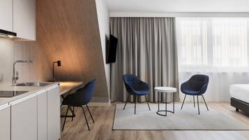 A modern hotel room with a kitchen area, two blue chairs, a small round table, and a large window with curtains.