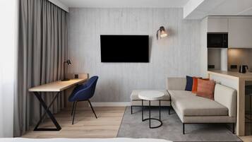 Una habitación de hotel moderna con un escritorio de madera, una silla azul, un sofá, una mesa redonda y un televisor de pantalla plana montado en la pared.