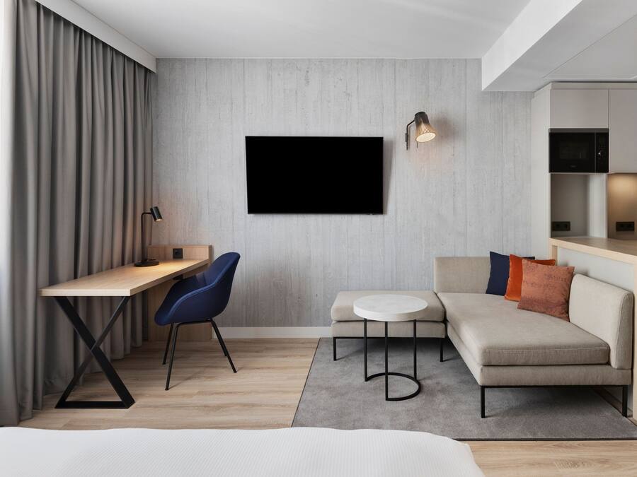 Una habitación de hotel moderna con un escritorio de madera, una silla azul, un sofá, una mesa redonda y un televisor de pantalla plana montado en la pared.