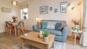 Ein Wohnzimmer mit blauem Sofa, Couchtisch aus Holz und einem Essbereich mit Stühlen und einem Tisch für zwei Personen.