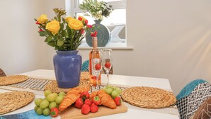 Ein Esstisch mit einer blauen Vase voller gelber und roter Blumen, einer Flasche Gallo-Wein und einem Glas mit einer Erdbeere. Auf dem Tisch liegen Trauben, Erdbeeren und Croissants.