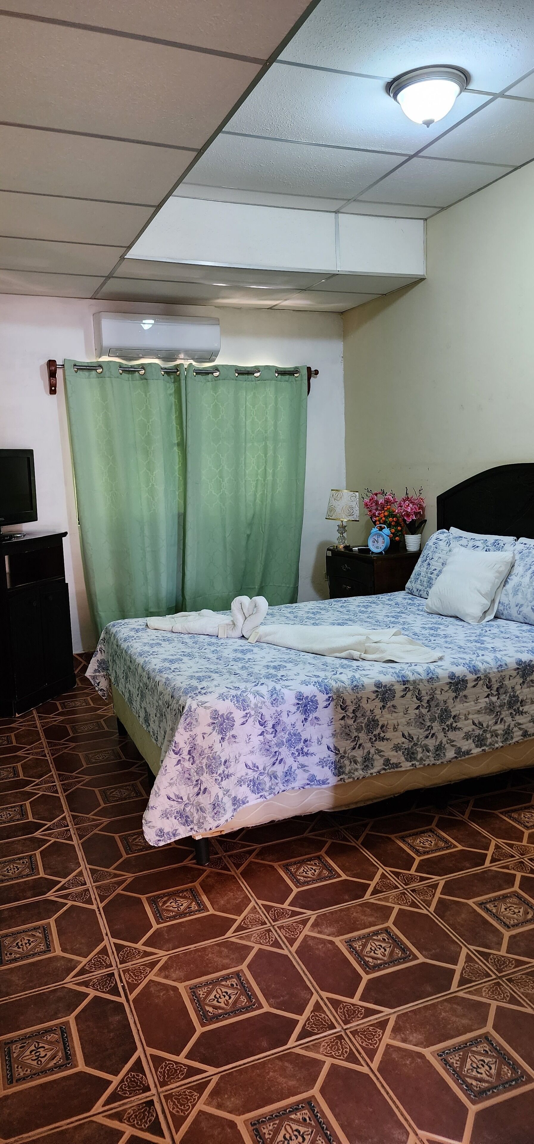 A hotel room with a bed, green curtains, a nightstand with a lamp and flowers, and a tiled floor.
