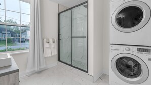A modern bathroom with a glass shower enclosure, a double washer-dryer unit, a large window with a view of the street, and a marble-tiled floor.