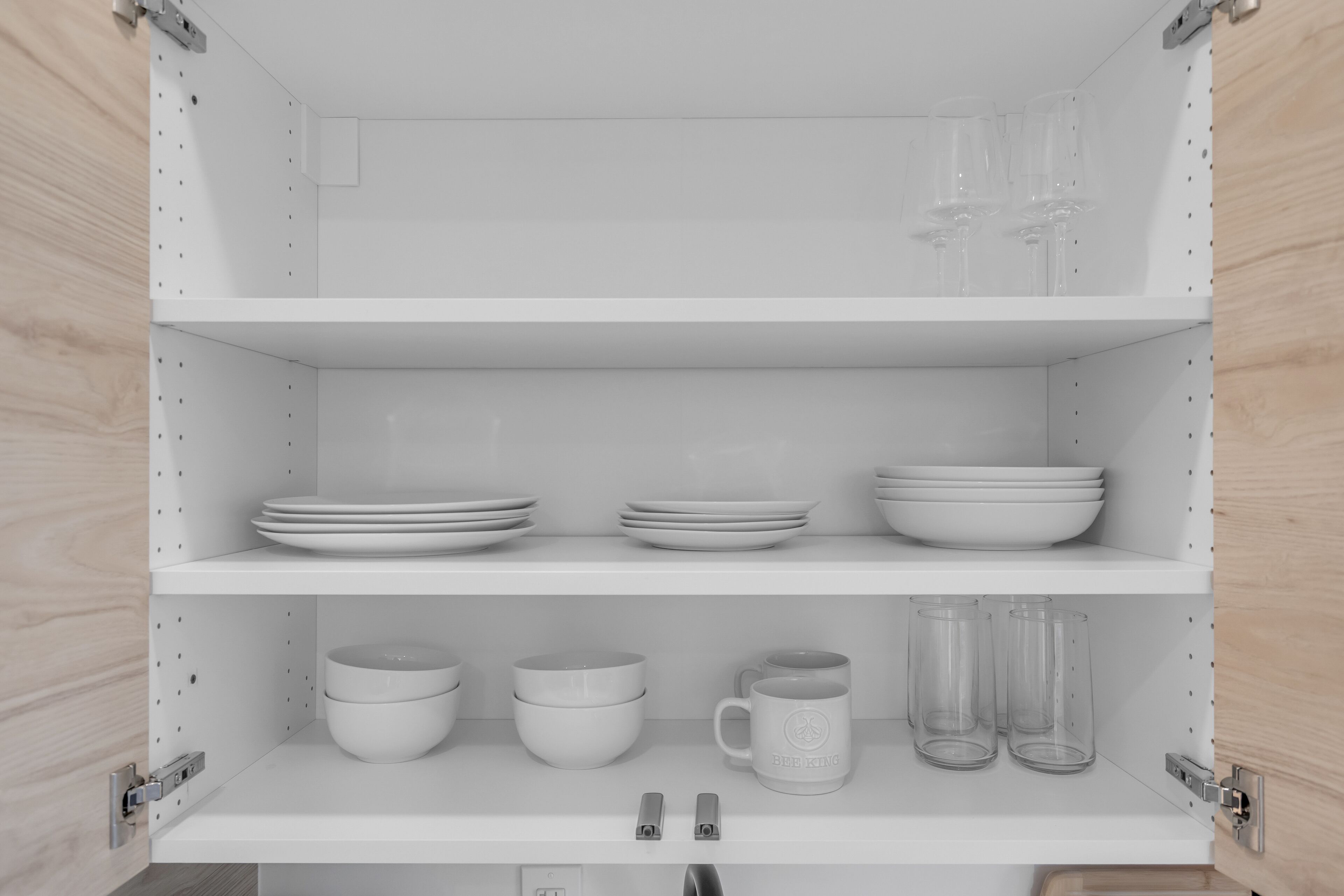 A white metal shelf unit with three shelves, each holding various white ceramic dishes and glasses.