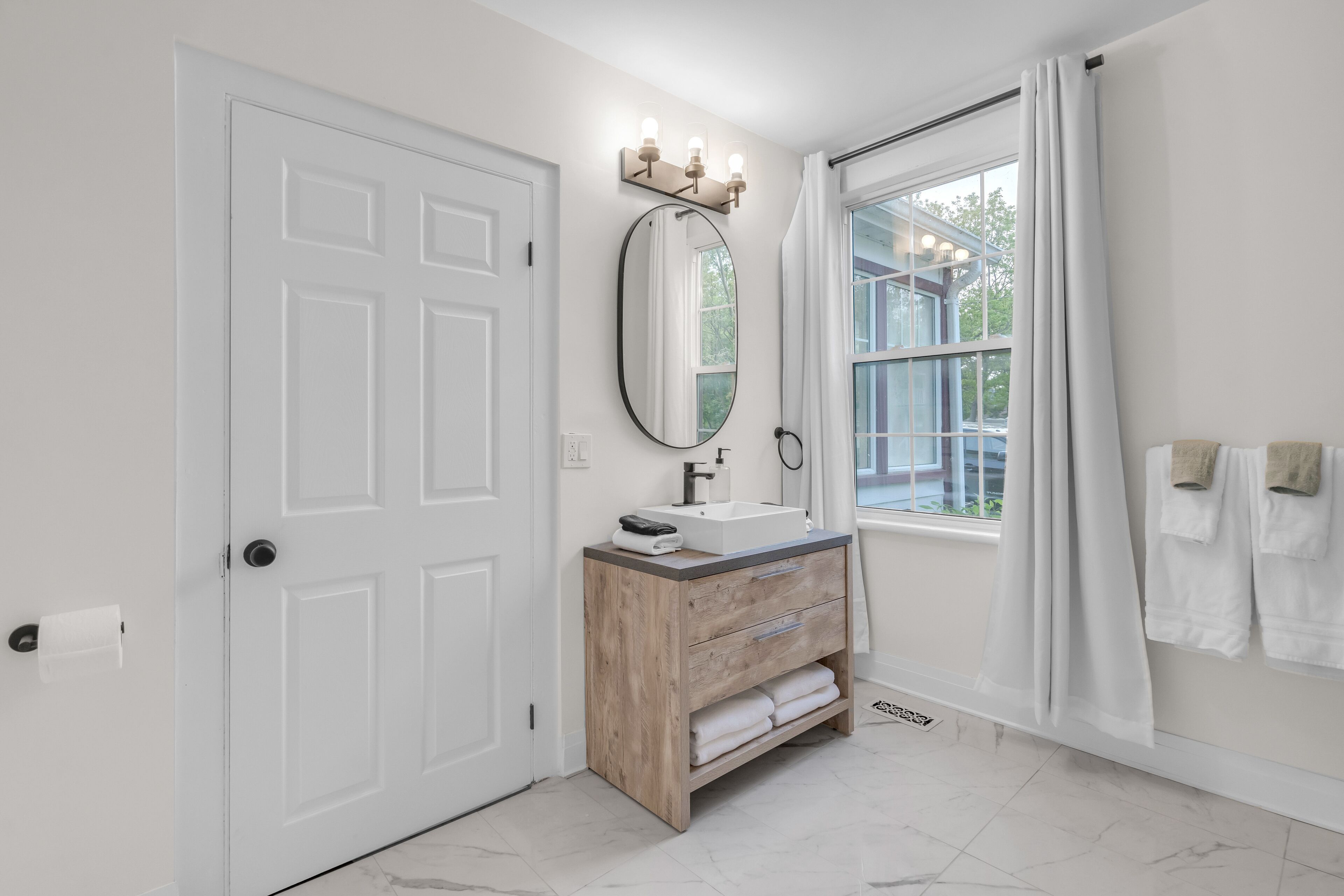 A bathroom with a white door, a wooden vanity with a mirror and lights, a window with curtains, and a towel rack with towels.