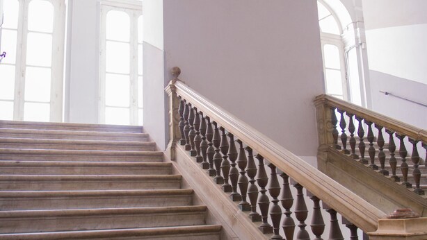 Een statige trap met een balustrade, die leidt naar een kamer met hoge plafonds en rondboogvensters.