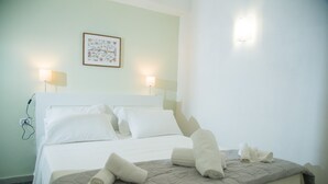 Een net opgemaakt bed met witte lakens en opgerolde handdoeken, een lijst met bloemkunst aan de muur, en twee bedlampjes.