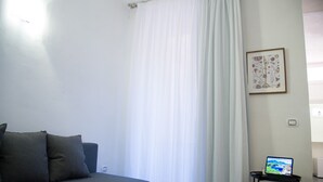 Een kamer met een grijze bank, een wit gordijn, een ingelijst schilderij aan de muur en een laptop op een klein tafeltje.
