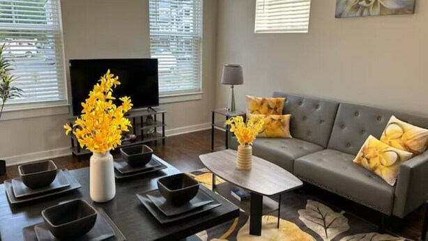 A modern living room with a grey sofa, a coffee table, and a TV. There are decorative vases with flowers and a painting on the wall. - Serenity 1 King Bed/Pool (Moseley)