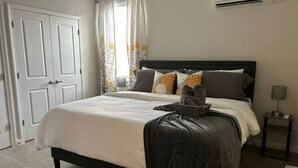 A bedroom with a bed, a nightstand, a lamp, a window with curtains, and an air conditioning unit. - Serenity 1 King Bed/Pool (Moseley)