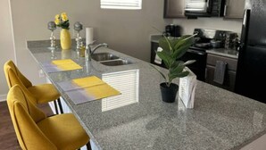 A kitchen with granite countertops, a sink, a stove, and cabinets. There are two yellow chairs and a potted plant on the counter. - Serenity 1 King Bed/Pool (Moseley)